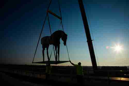 A horse sculpture on a crane is silhouetted against the sun, which is low in the sky. Two people steady the platform the horse is standing on.